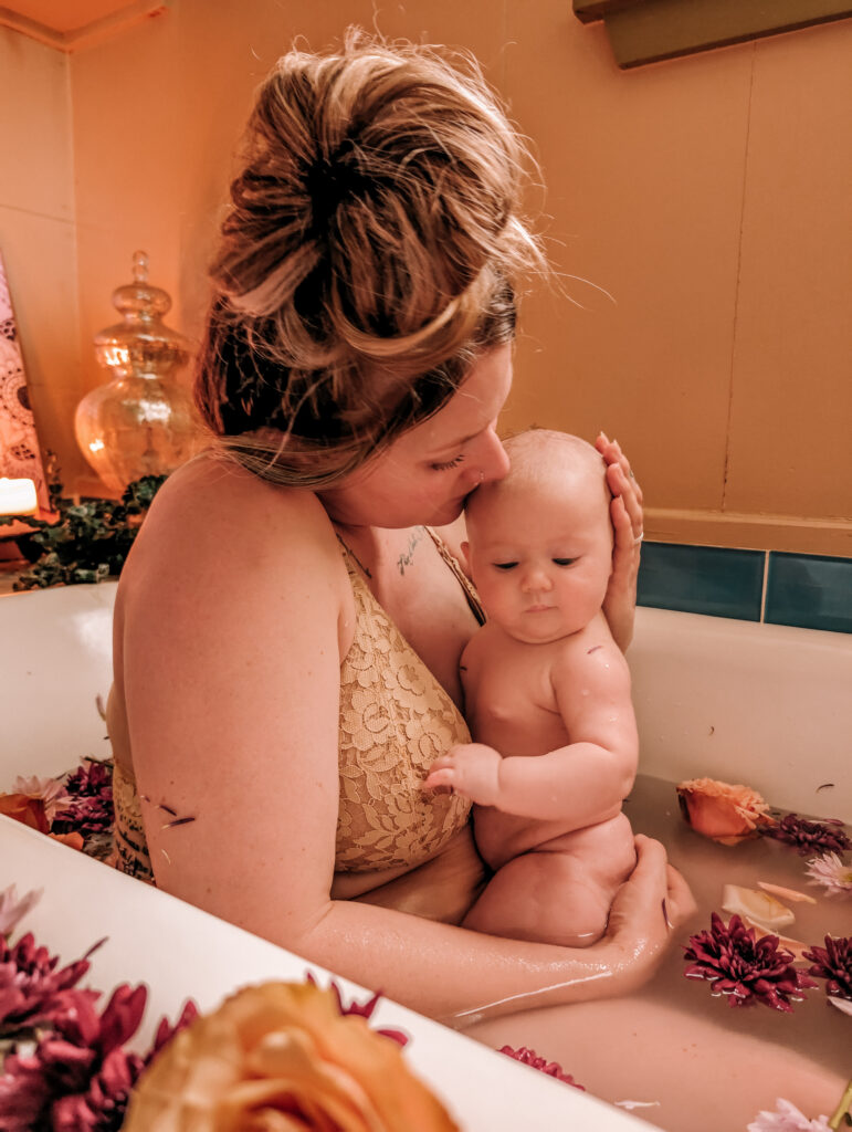 Mother and baby posing in an embrace for the camera while sitting in a bath filled with flowers and petals for a Rebirthing Ritual