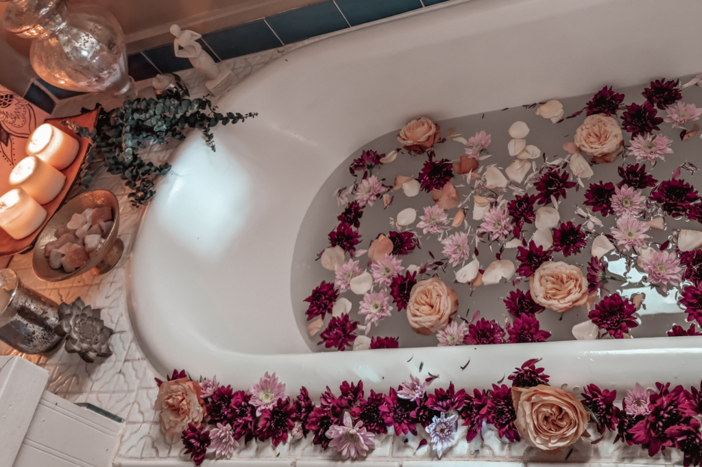 White claw foot bath tub filled with milky water, garden roses, dahlias, and flower petals with cleansing crystals and herbs for a Rebirthing Ritual to reclaim a birth story